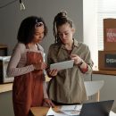 Young confident female entrepreneur pointing at screen of tablet with online information while explaining something to colleague